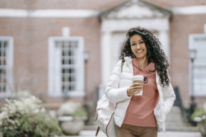 Cheerful young ethnic woman drinking takeaway coffee and smiling near brick building